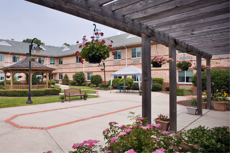 Outdoor courtyard area of a senior living facility with a wooden pergola, hanging flower baskets, a gazebo, benches, and a white canopy tent with chairs underneath. The building is two stories with brick walls and multiple windows.