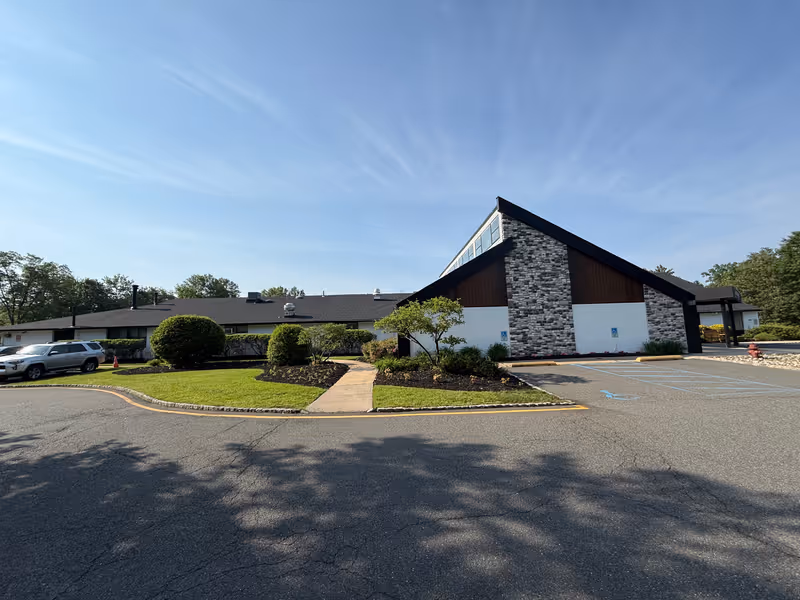 Front exterior of a single-story senior living facility with a triangular peaked entrance, parking lot, and landscaped lawn under a clear blue sky.