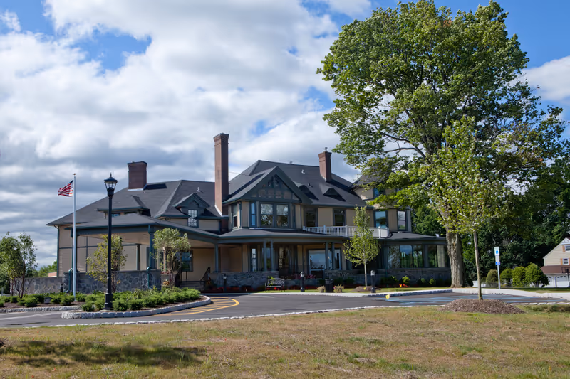Exterior view of a large, multi-story senior living facility building with a wraparound porch, several chimneys, and large windows. The building is surrounded by landscaped greenery, trees, a flagpole with an American flag, and a paved driveway with parking spaces. The sky is partly cloudy.