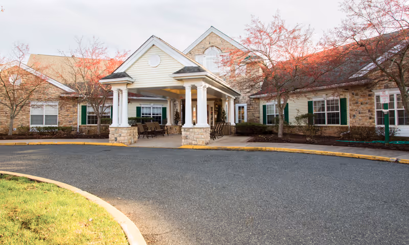 Front entrance of a brick-and-siding building with a covered porte-cochère, columns, seating, and a curved driveway.