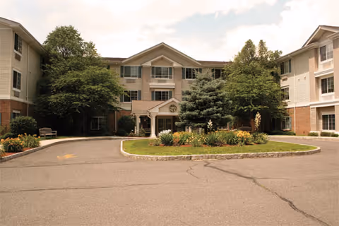 Front exterior view of a three-story senior living facility building with a circular driveway and landscaped garden area in the center. The building has beige siding with brick accents and multiple windows. Trees and shrubs surround the entrance area.