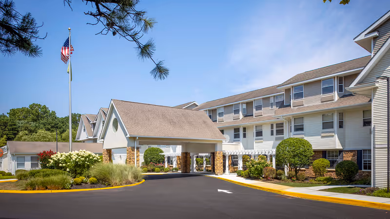 Exterior view of a senior living facility with a covered entrance, landscaped bushes and trees, and an American flag on a flagpole against a clear blue sky.