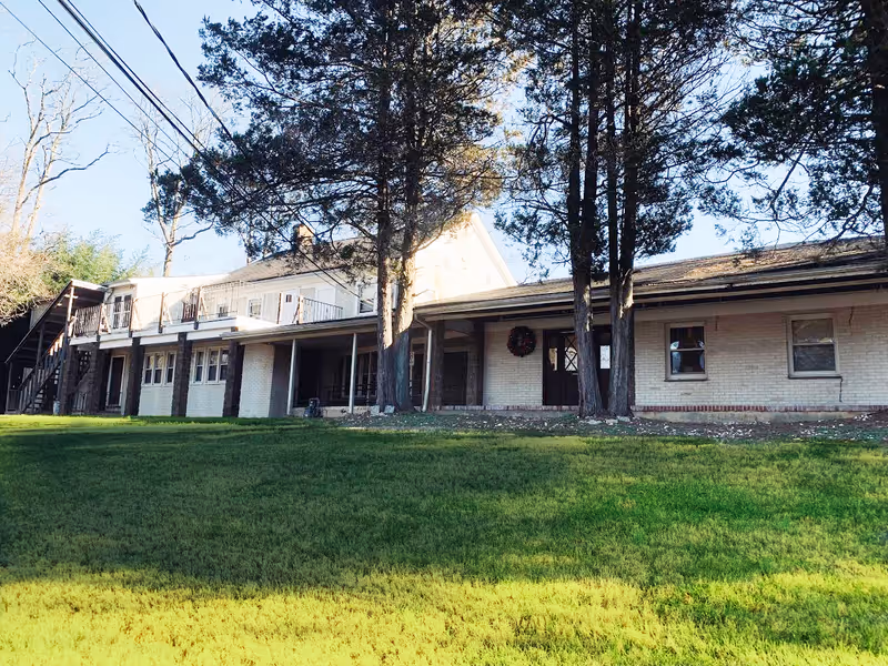 Exterior front view of a two-story brick building with a covered porch, trees, and a grassy lawn.