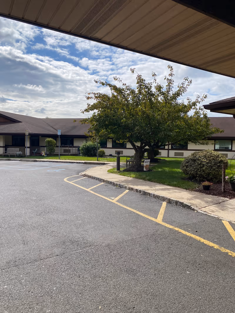 View of the front courtyard and parking area of a single-story senior living facility with a tree, walkway, and building in the background.