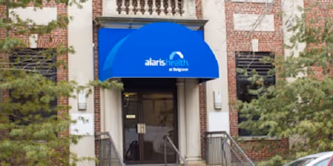 Entrance to a brick building with a blue awning that reads 'alaris health at Belgrove' above glass double doors. There are stairs with metal railings leading up to the entrance, and some greenery is visible on the left side.