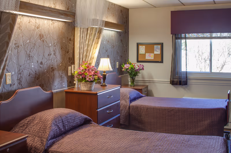 A cozy bedroom with two single beds covered in purple bedspreads. Between the beds is a wooden nightstand with a lamp and a basket of flowers. Another small table with a vase of flowers is near the window, which has sheer and purple curtains. The walls are decorated with patterned wallpaper and a bulletin board with pinned notes.