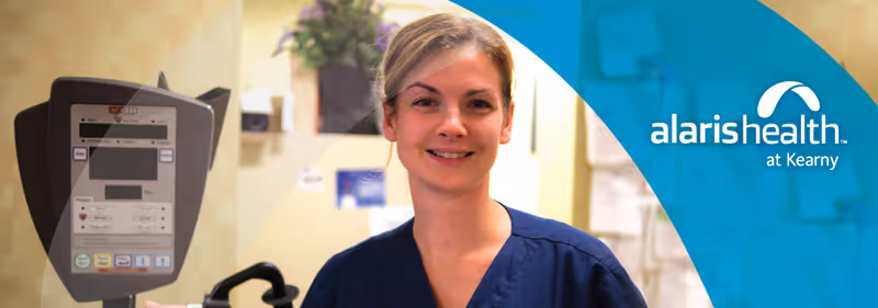 A smiling healthcare worker in blue scrubs standing in a medical facility with medical equipment and a plant in the background. The image includes the logo and name 'alaris health at Kearny' on a blue overlay on the right side.
