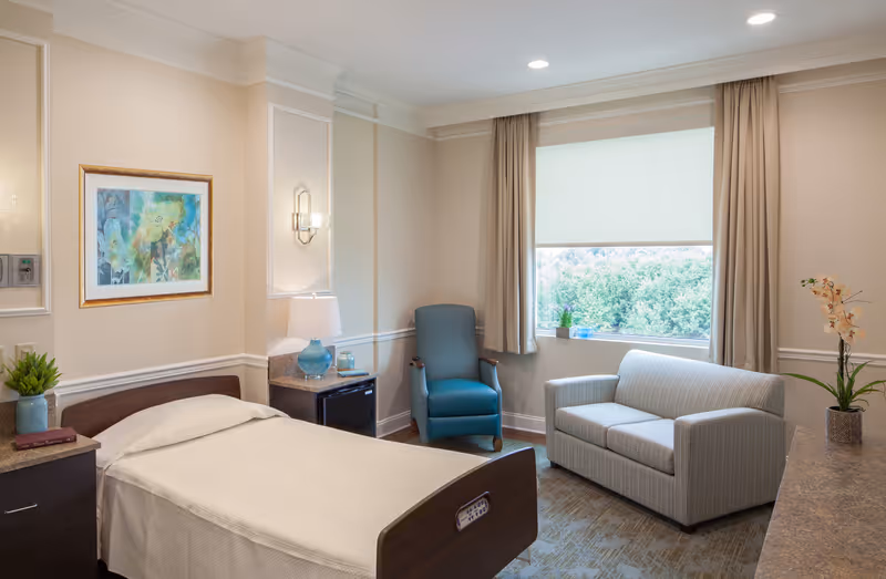 A well-lit bedroom in a senior living facility featuring a single bed with white bedding, a wooden headboard, a nightstand with a blue lamp and a small plant, a comfortable blue armchair, a striped loveseat, and a large window with beige curtains overlooking greenery outside. The walls are light-colored with decorative molding and a framed abstract painting.