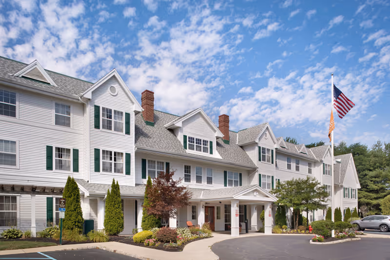 Exterior view of a large, white senior living facility building with green shutters and multiple windows under a partly cloudy blue sky. The entrance has a covered porch with columns, surrounded by landscaped bushes and trees. An American flag and another flag are flying on a flagpole near the entrance. Several cars are parked in the parking lot in front of the building.