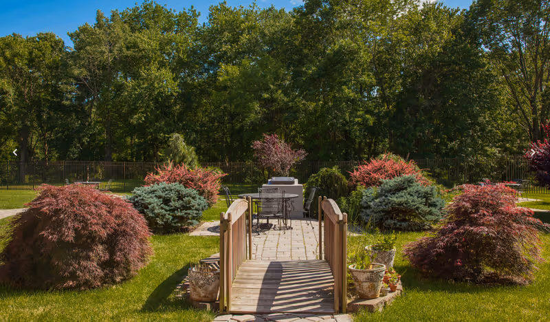 A landscaped outdoor courtyard with a small wooden footbridge leading to a patio table and chairs surrounded by shrubs and trees.