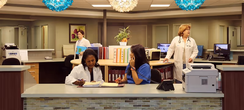 A medical office reception area with two women seated at the front desk, one writing in a binder and the other talking on the phone. Two other women in white lab coats are standing in the background near desks and computers. The area is well-lit with decorative blue and white hanging lights and has organized files and office equipment.