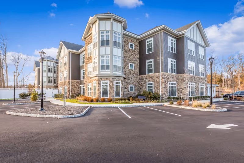 Exterior view of a three-story senior living facility building with stone and siding facade, multiple windows, and a parking lot in front under a blue sky with some clouds.