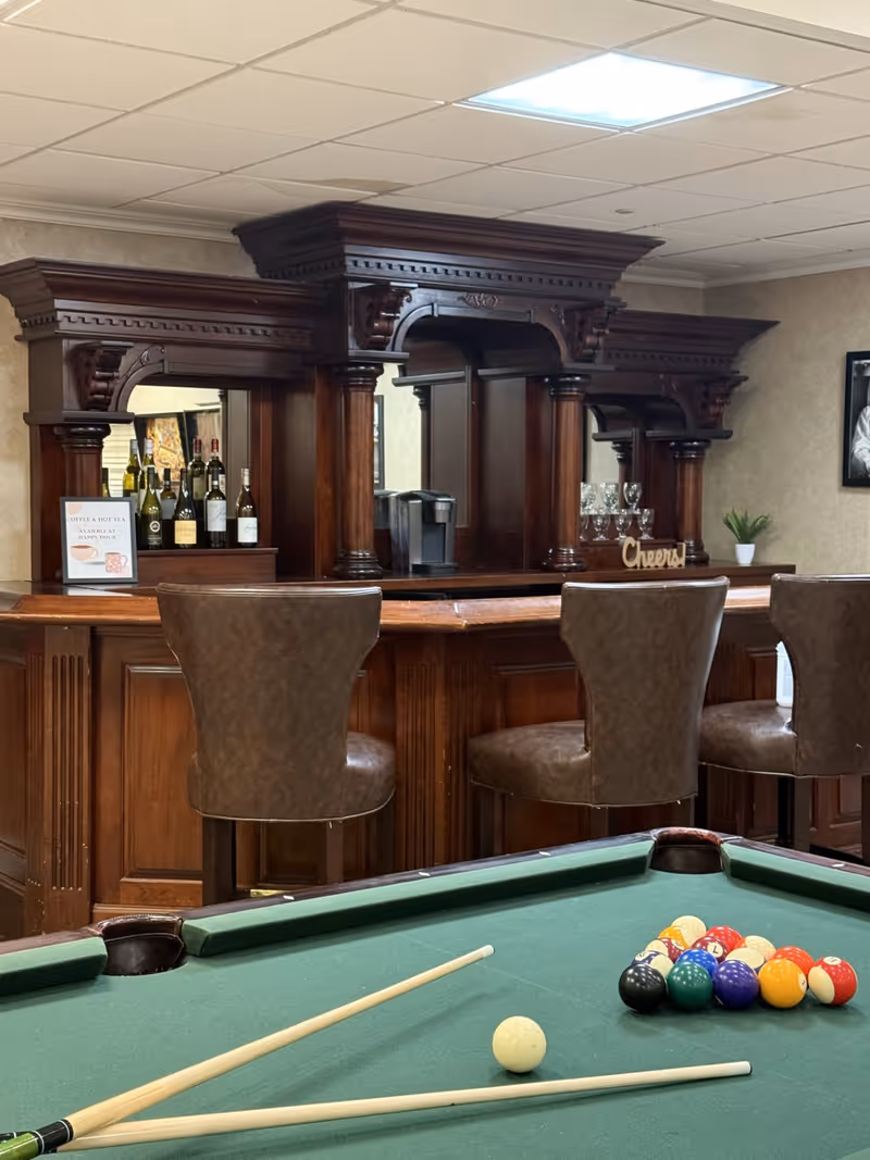 Interior view of a recreational room featuring a green pool table with billiard balls and cues in the foreground. Behind the pool table is a wooden bar with three brown leather bar stools. The bar has an ornate wooden back with shelves holding bottles of wine and glasses. A small sign on the bar reads 'Coffee & Hot Tea Available'.