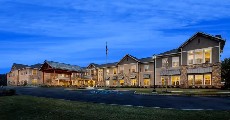 Exterior view of a two-story senior living facility building at dusk with lights glowing from the windows, a covered entrance with wooden beams, an American flag on a flagpole, and a paved driveway with parking spaces in front.