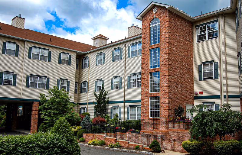 Exterior view of a multi-story assisted living facility with beige siding and a prominent brick tower featuring large windows. The building is surrounded by well-maintained landscaping including bushes, trees, and flowers under a partly cloudy blue sky.
