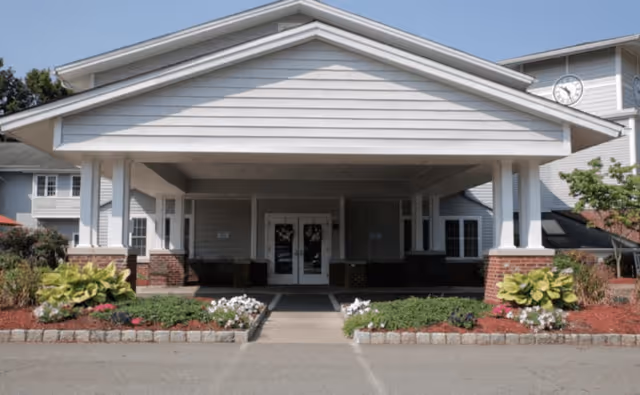 Front exterior view of a building with a covered entrance supported by white columns. There are flower beds with green plants and white flowers on either side of the walkway leading to double glass doors. The building has light-colored siding and a clock is visible on the upper right side.
