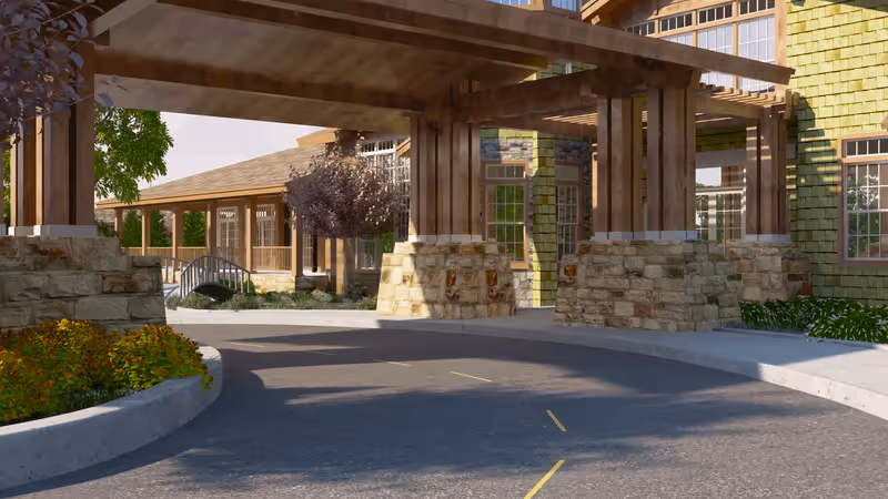 Covered driveway entrance to a building with wooden beams and stone pillars, surrounded by landscaped greenery and trees, with a pathway and a small bridge visible in the background.
