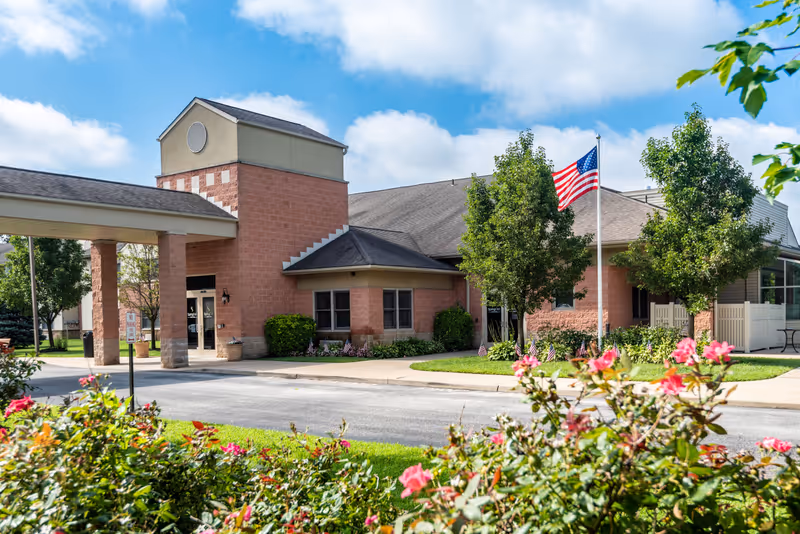 Exterior view of Spring Oak at Berlin facility showing a brick building with a covered entrance, an American flag on a flagpole, green trees, and blooming pink flowers in the foreground under a partly cloudy blue sky.