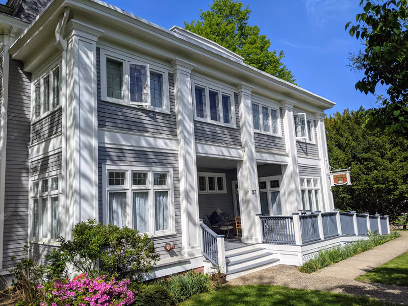 Exterior view of a two-story building with gray siding and white trim, featuring multiple windows and a front porch with railings. There is a sign that reads 'Montclair Inn' hanging near the entrance. The building is surrounded by green trees, bushes, and a lawn with a concrete walkway.