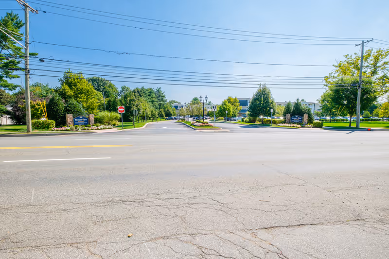 Street view across a road showing the landscaped entrance and sign for Brightview Paramus senior living with buildings and trees behind.