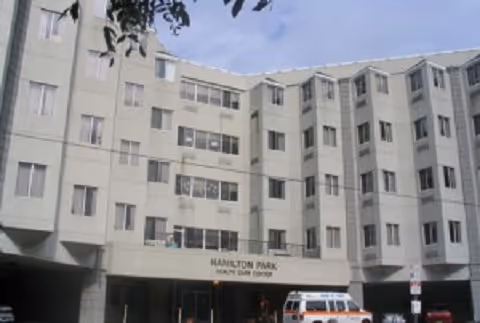 Exterior view of a multi-story senior care facility building with multiple windows and a covered entrance. An ambulance is parked in front of the entrance, and there are some trees partially visible at the top.