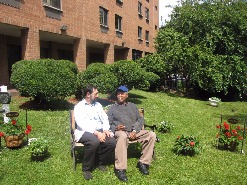 Two men sitting and talking on a wooden bench in a grassy garden area with neatly trimmed bushes and flowering plants, adjacent to a brick building.
