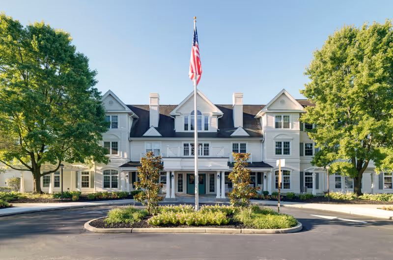 Front exterior view of a large, white, multi-story senior living facility building with a central entrance, surrounded by green trees and landscaping. An American flag is prominently displayed on a flagpole in the center of a circular driveway.