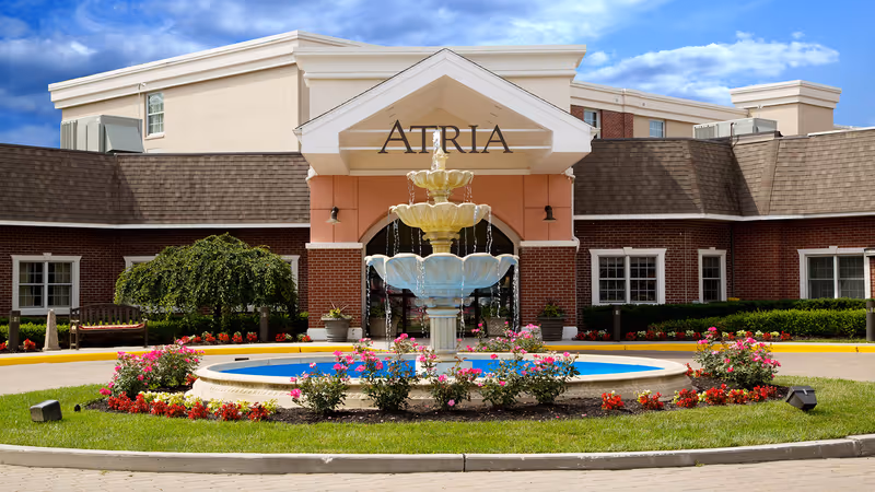 Front exterior view of Atria Cranford senior living facility featuring a large three-tiered fountain surrounded by colorful flowers and greenery, with a brick building and entrance canopy displaying the name 'Atria' under a blue sky with some clouds.