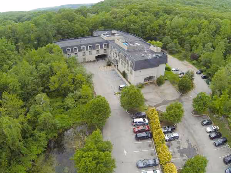 Aerial view of a large building surrounded by dense green trees and a parking lot with several cars parked. The building has a flat roof and multiple windows, situated in a forested area.