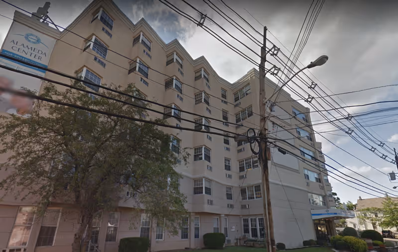 Multi-story beige Alameda Center for Rehabilitation and Healthcare building seen from the street with power lines and a tree in front.