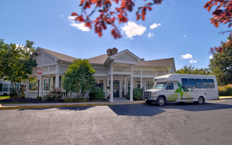 Exterior view of The Residence at Stafford building with a covered entrance, surrounded by trees and bushes under a clear blue sky. A white shuttle bus with green leaf graphics and the facility's name is parked in front.