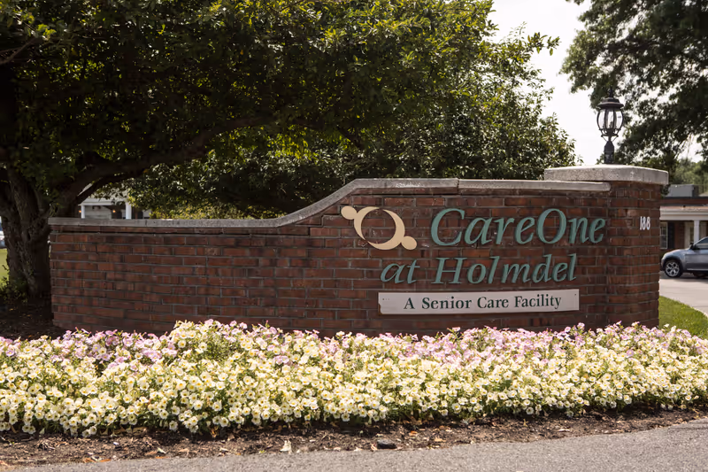 A brick sign at the entrance of CareOne at Holmdel, a senior care facility, surrounded by blooming white and light pink flowers with a large tree in the background.