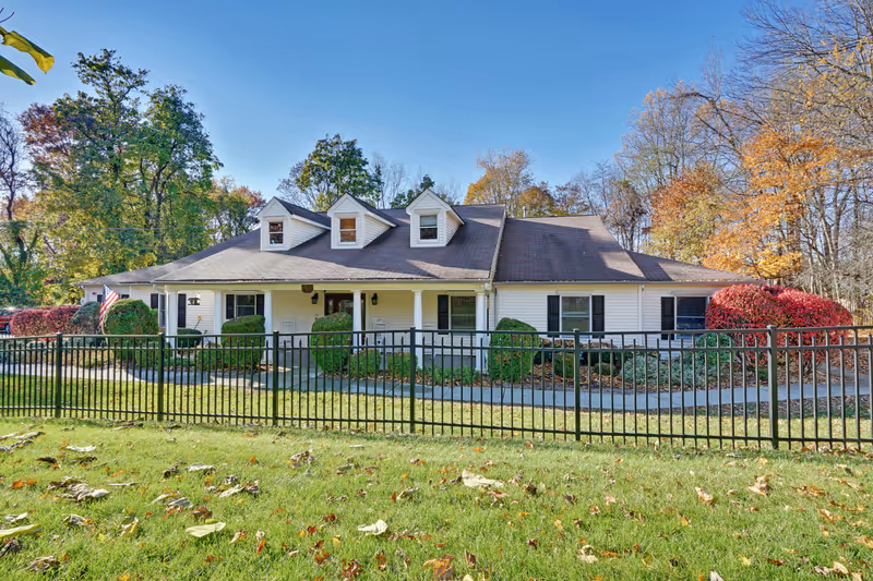 Single-story white building with a dark roof and three dormer windows, surrounded by a black metal fence and landscaped bushes. Trees with autumn foliage are visible in the background under a clear blue sky.