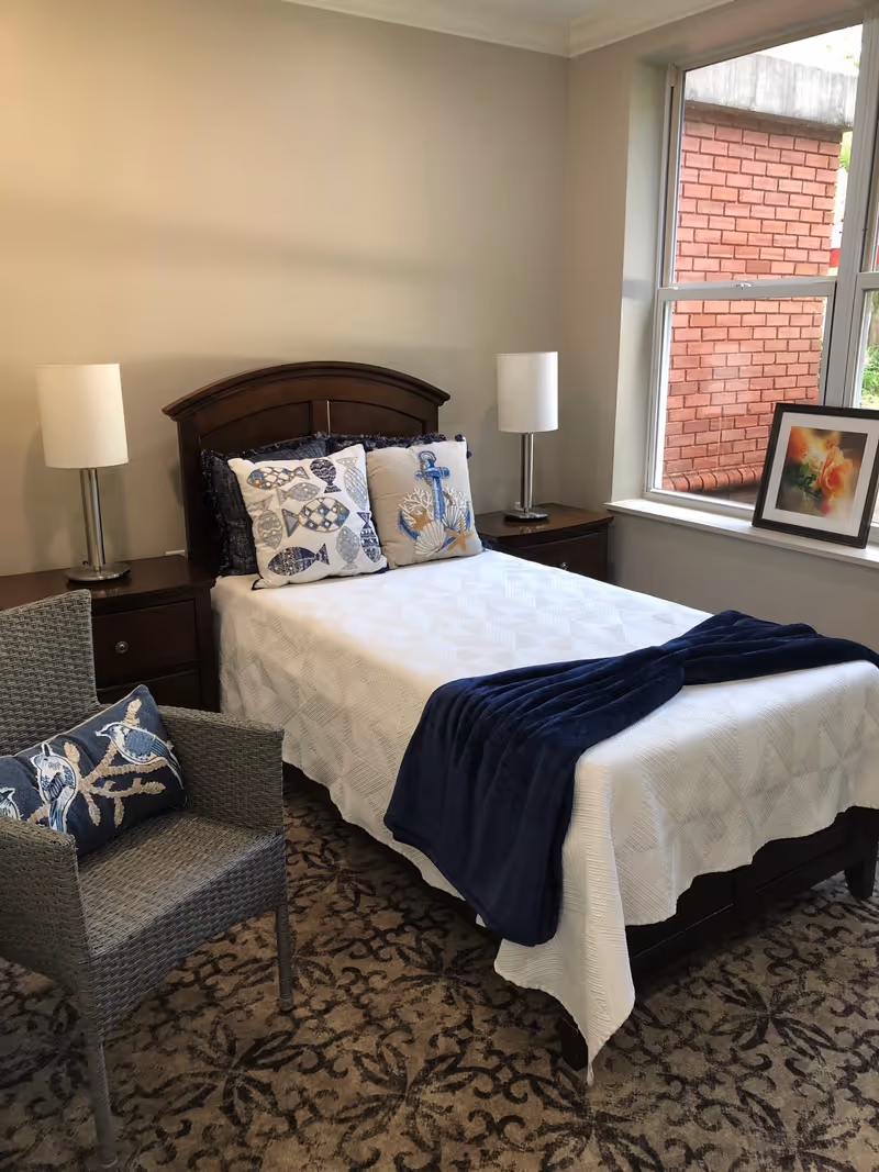 A neatly made single bed with a white textured bedspread and a dark blue throw blanket at the foot. The bed has a dark wooden headboard and is flanked by two matching wooden nightstands, each with a modern white lamp. There are two decorative pillows with fish and nautical designs on the bed. A wicker chair with a blue and white pillow is positioned next to the bed. A window with a view of a brick wall lets in natural light, and a framed floral painting rests on the windowsill. The floor is covered with a patterned carpet.