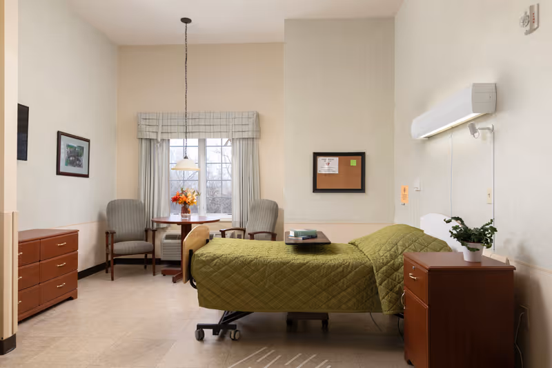 A senior living facility bedroom with a single bed covered in a green quilt, a wooden nightstand with a potted plant, a small table with two upholstered chairs near a window with curtains, a dresser, and a bulletin board on the wall.