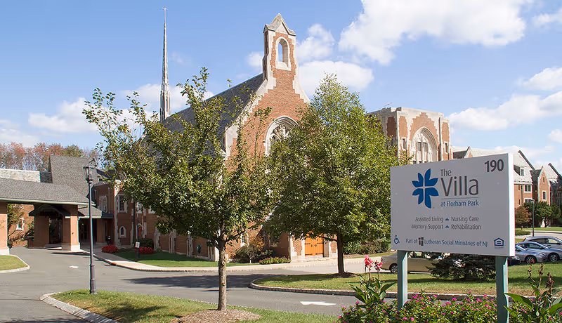 Exterior view of The Villa at Florham Park senior living facility showing a large brick building with gothic architectural elements, a tall spire, trees, a driveway, and a sign with the facility's name and services offered.