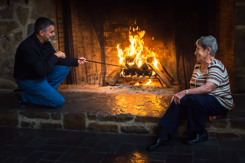An elderly woman sitting on a stone hearth smiling at a man who is crouched down tending to a fire in a large stone fireplace.