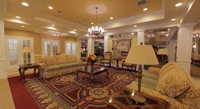 A spacious and elegantly decorated living room with patterned sofas, a large ornate rug, wooden coffee tables, table lamps, and a chandelier hanging from the ceiling. The room features cream-colored walls, columns, and multiple doorways with glass panels.