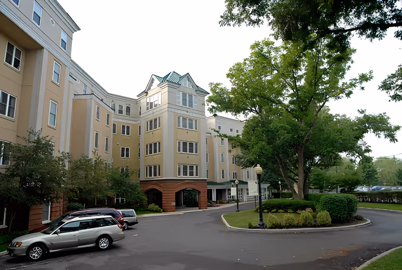 Exterior front view of a multi-story senior living building with a circular driveway, parked cars, and landscaped trees.