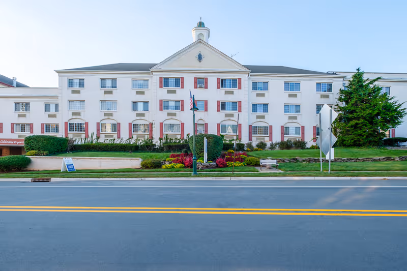 Front exterior of a three-story white senior living building with red shutters, landscaped lawn and a street in the foreground.