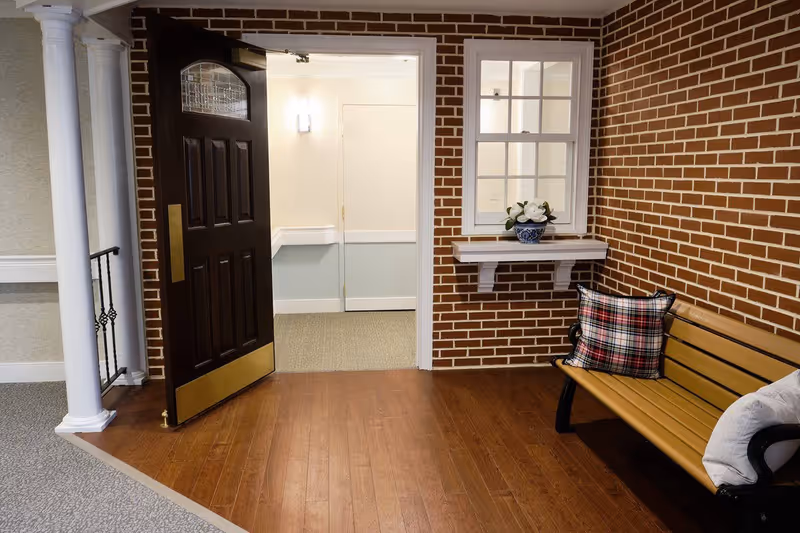 An open dark wooden door with a brass kick plate opens into a hallway with light-colored walls and carpet. To the right of the door is a brick wall with a small white-framed window above a white shelf holding a blue and white vase with white flowers. Below the window is a wooden bench with black metal armrests, decorated with a plaid cushion and a white pillow. The floor near the door is wooden, transitioning to carpet further inside. Two white columns are visible on the left side of the image.