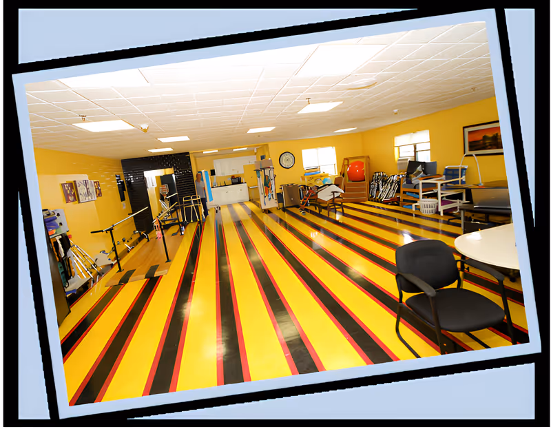 A spacious room with bright yellow walls and a floor featuring bold yellow, black, and red stripes. The room contains various exercise and therapy equipment, including parallel bars, chairs, and other physical therapy tools. There are windows letting in natural light, a clock on the wall, and some framed artwork.