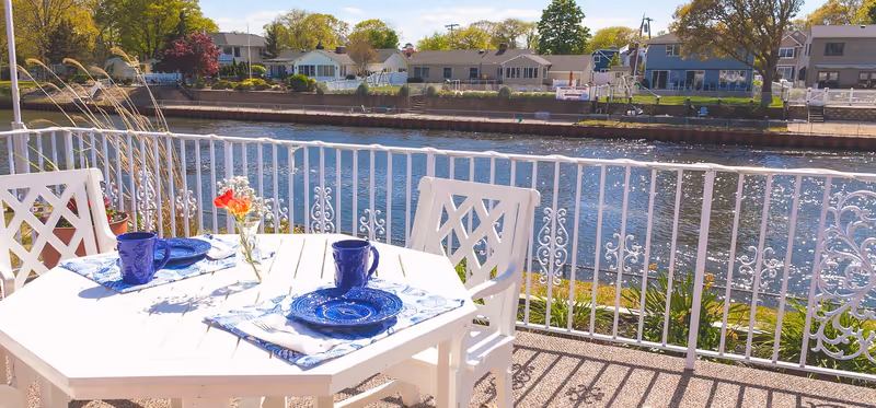 Outdoor patio area with a white table and two white chairs set with blue plates and mugs, overlooking a calm river with houses and trees on the opposite bank under a clear sky.