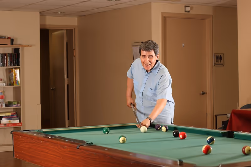 An elderly man playing pool indoors, leaning over a pool table and aiming with a cue stick. The room has beige walls, a bookshelf with board games and books, and a door with a restroom sign in the background.