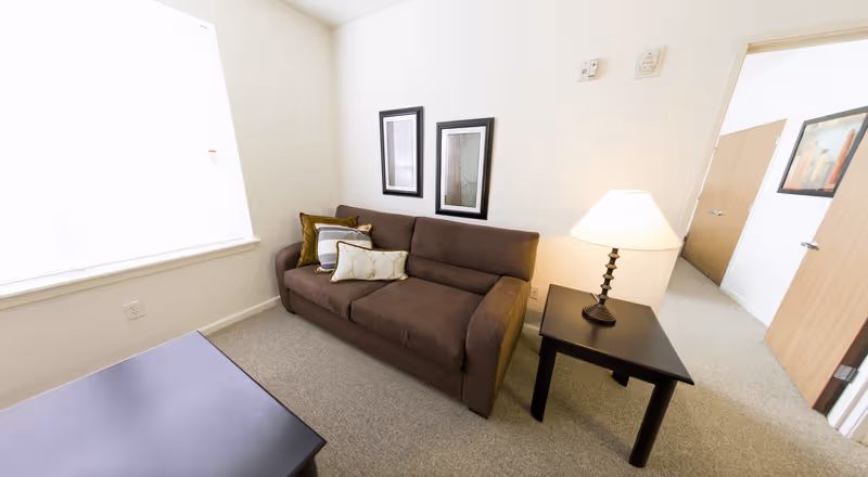 Small living room with a brown sofa, side table and lamp, coffee table, bright window, and framed wall art.