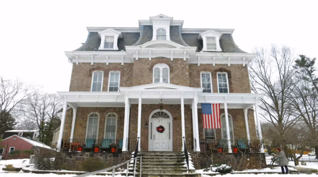 Front exterior view of a large, historic stone building with white trim and a porch. The building has multiple windows, a central door with a wreath, an American flag hanging on the right side of the porch, and snow on the ground. A person is visible near the right side of the image.