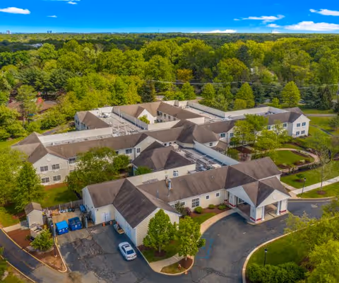 Aerial view of Spring Hills Somerset senior living facility surrounded by lush green trees and landscaping under a bright blue sky with scattered clouds. The building has multiple connected sections with beige walls and brown roofs, and a parking area with a few vehicles is visible in front.