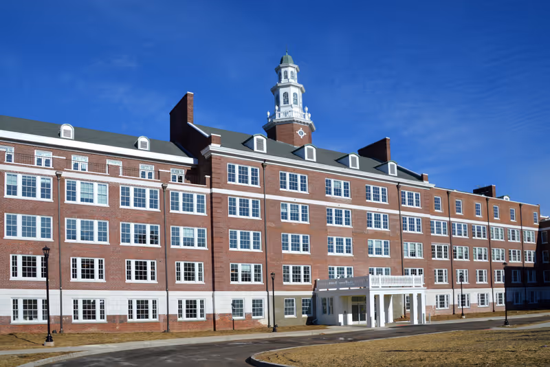Exterior view of a large multi-story brick building with many windows and a white cupola on the roof, under a clear blue sky.