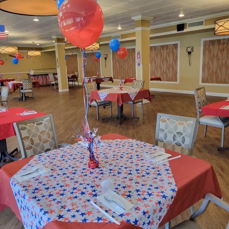 A dining room decorated with red, white, and blue star-themed tablecloths and balloons. Several tables are set with napkins and utensils, and the room has wooden flooring, beige walls, and patterned curtains. The atmosphere suggests a festive or patriotic celebration.