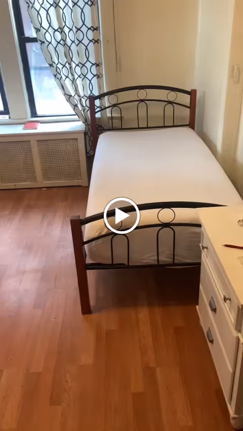 A small bedroom with a single bed featuring a metal and wood frame, a white mattress, a white dresser with drawers, and a window with patterned curtains letting in natural light.